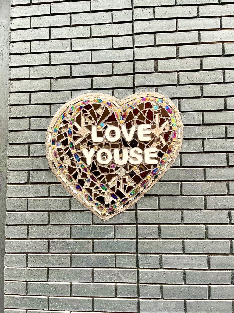 A multi-colored mosaic heart by the Philly-basedartist Amberella with the words Love Youse written with while tiled letters in the center of the heart. The mosaic is on a white brick wall with black grouting.